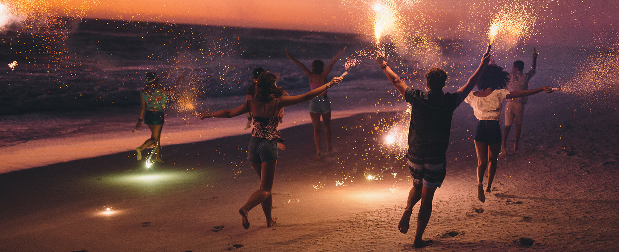 A group of young adults running on the beach with sparklers by the beach.